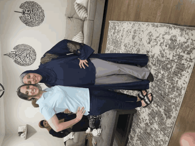two women standing next to each other in a living room with arabic writing on the wall