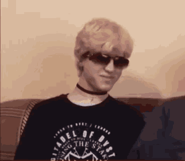 a young man wearing sunglasses and a choker is sitting on a couch and smiling .