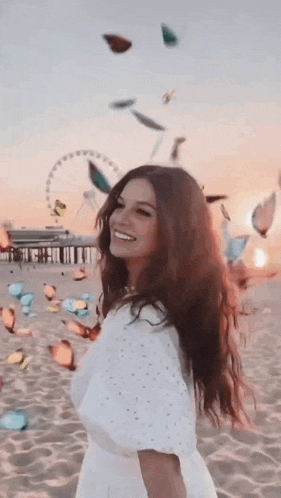 a woman in a white dress is standing on a beach with butterflies falling around her .