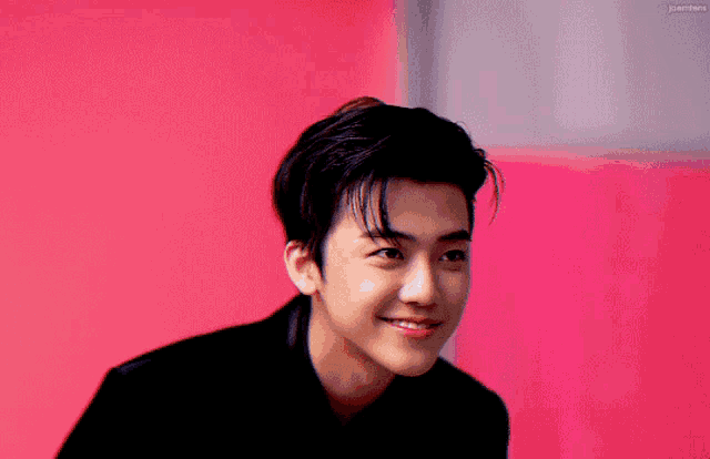 a young man in a black suit is smiling in front of a pink background .