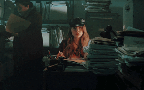 a woman wearing a black hat sits at a desk with a stack of papers