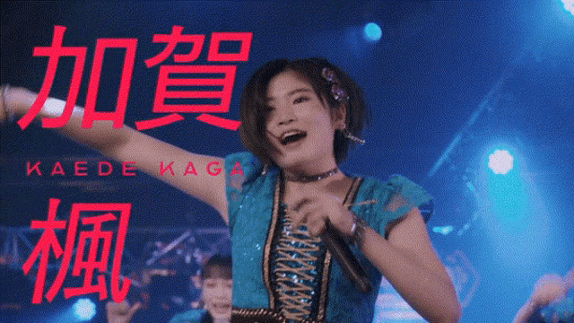 a woman in a blue dress singing into a microphone with kaede kaga in red letters behind her