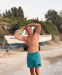 a shirtless man in blue swim trunks is standing on a beach next to a boat .