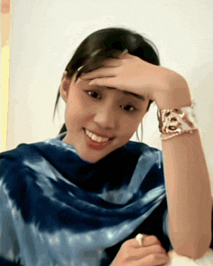 a woman wearing a blue scarf holds her hand to her forehead and smiles