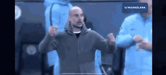 a man in a gray jacket is standing on a soccer field with his hands in the air .