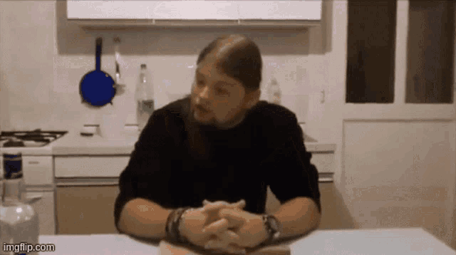 a man with long hair is sitting at a table with his hands folded in front of a kitchen counter