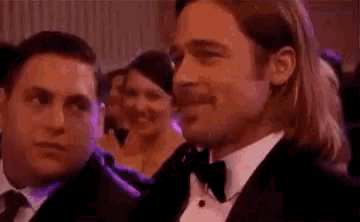 a man in a tuxedo is smiling while sitting in a crowd of people at a party .