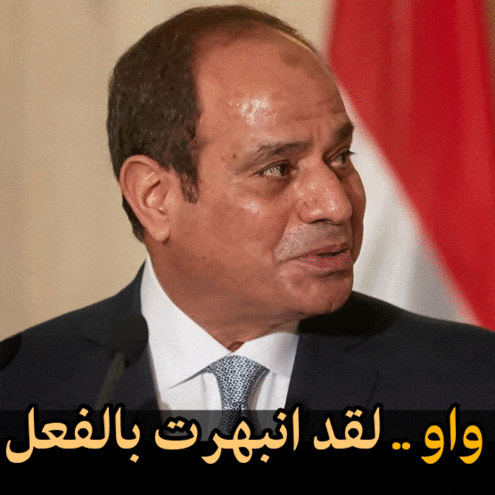 a man in a suit and tie stands in front of a microphone with arabic writing behind him