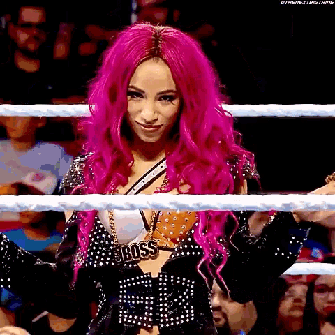 a woman with pink hair is standing in a wrestling ring wearing a boss necklace .