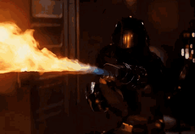 a man in a helmet is holding a flamethrower in front of a fire in a dark room .