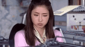 a woman is sitting at a desk with a scarf around her neck and making a funny face .