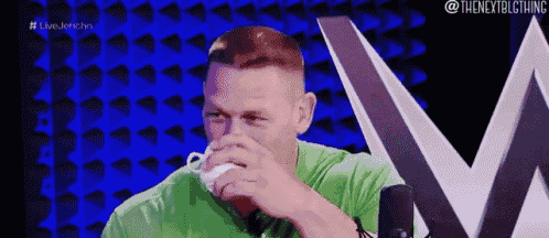 a man in a green shirt is sitting in front of a microphone and drinking from a white cup .