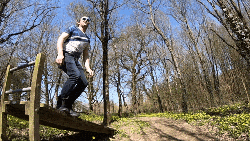 a man wearing sunglasses stands on a wooden bridge in the woods