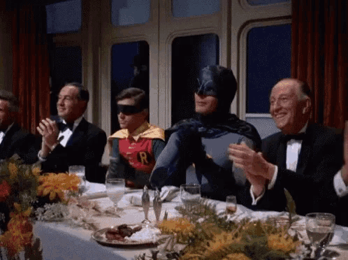 a group of men are sitting at a table with batman and robin applauding