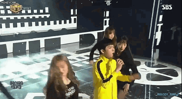 a man in a yellow jacket is singing into a microphone on a stage surrounded by women .