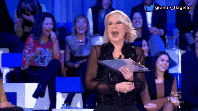 a woman is laughing while holding a clipboard in front of a crowd with the hashtag grande flagello at the top