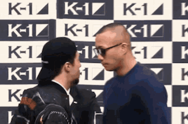 two men are standing next to each other and looking at each other in front of a sign that says k-1 .
