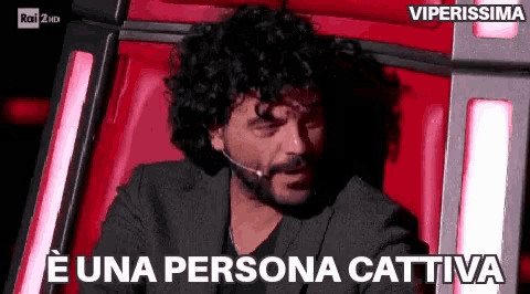 a man with curly hair and a beard is sitting in a red chair with a microphone in his mouth .