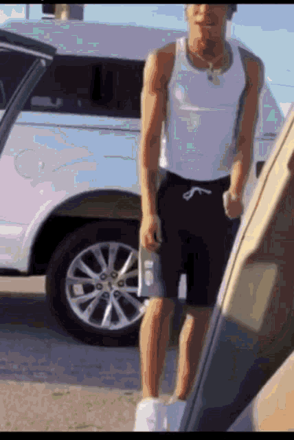 a man in a white tank top and black shorts is standing next to a car