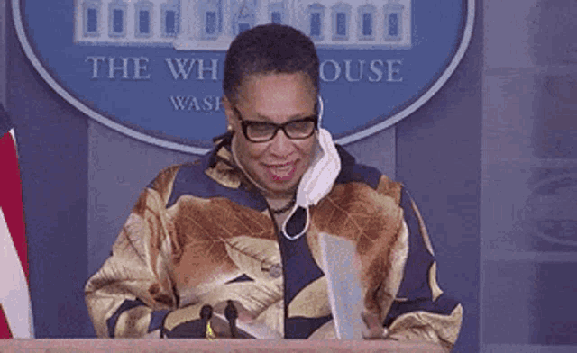 a woman wearing glasses and a mask is sitting at a podium in front of a sign that says the white house