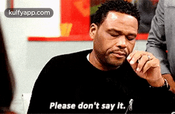 a man in a black sweater is sitting at a table and says please don 't say it .