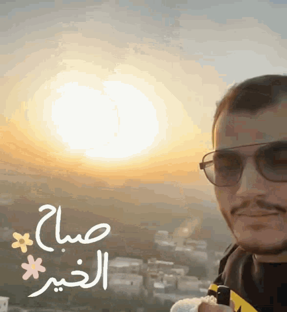 a man wearing sunglasses stands in front of a sunset with the words " صباح الخير " written in arabic