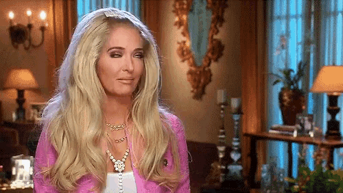 a blonde woman is sitting in a living room wearing a pink jacket and a pearl necklace .