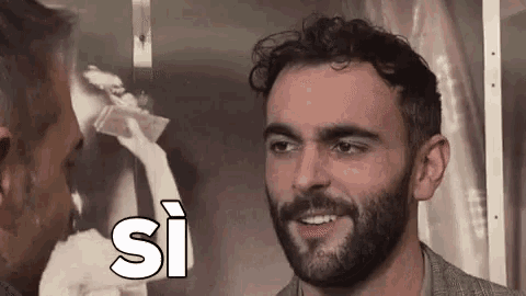 a man with a beard is smiling and talking to another man in front of a picture of a woman .