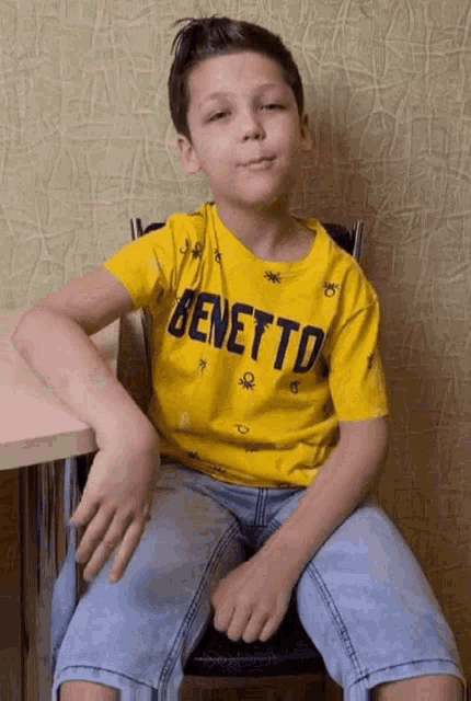 a young boy is sitting in a chair wearing a yellow t-shirt that says benetton .