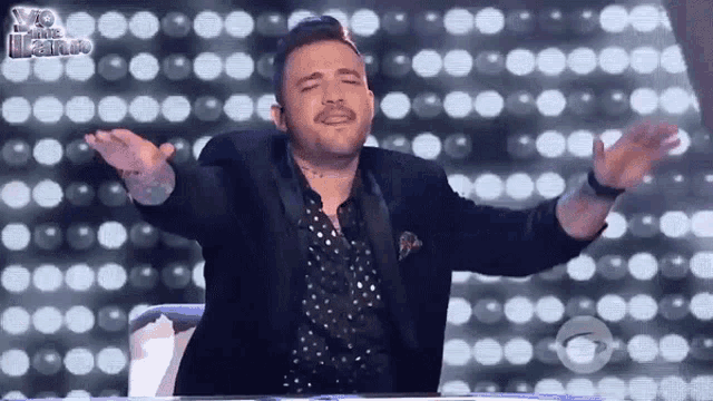 a man in a suit and polka dot shirt is sitting in front of a wall of lights with his arms outstretched .
