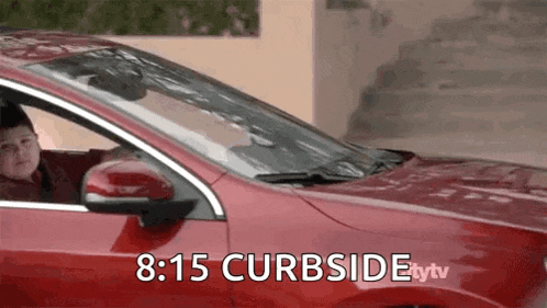 a young boy is driving a red car with the time of 8:15 curbside