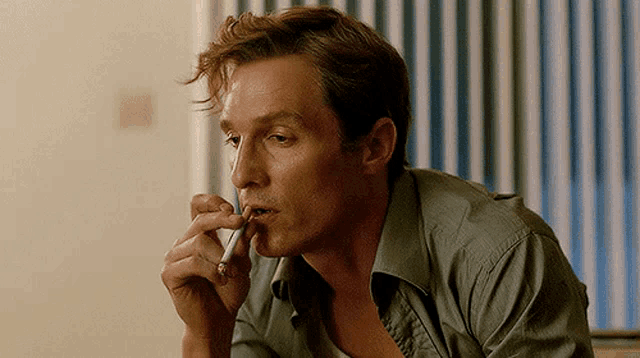 a man in a green shirt is smoking a cigar in front of a window