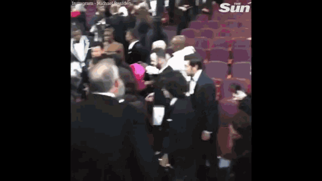 a crowd of people are sitting in a theater with the sun written on the bottom of the screen .