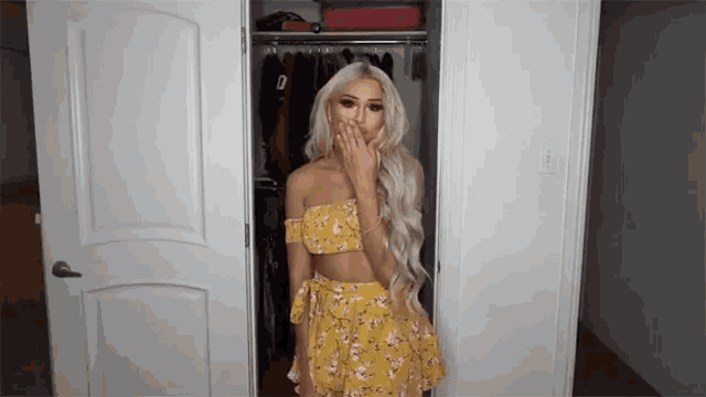 a woman in a yellow dress is standing in a closet and covering her mouth with her hand .