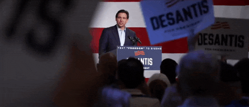 a man stands at a podium in front of a crowd holding a sign that says desantis