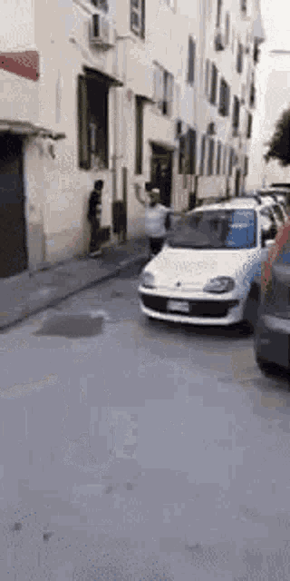 a white car is driving down a street next to a building with a man standing on the sidewalk .