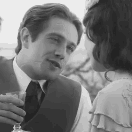 a man in a suit and tie is holding a wine glass and talking to a woman