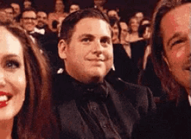 a man in a tuxedo is sitting in a crowd with a woman and a man .