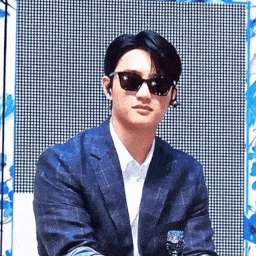 a man wearing sunglasses and a suit is sitting in front of a large screen .