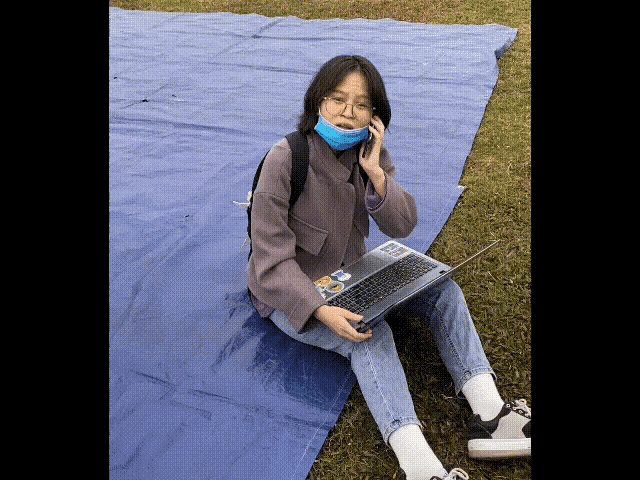 a girl wearing a mask is sitting on a blue tarp talking on a cell phone while holding a laptop