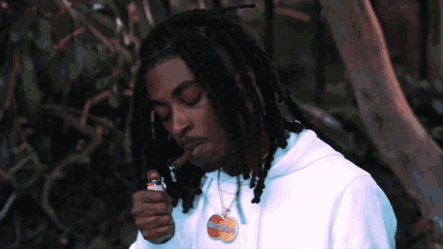 a man with dreadlocks is lighting a cigarette with a mastercard necklace around his neck