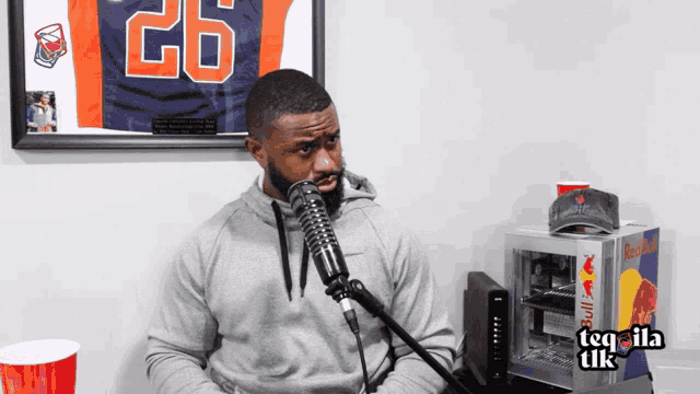 a man talking into a microphone in front of a framed jersey with the number 26