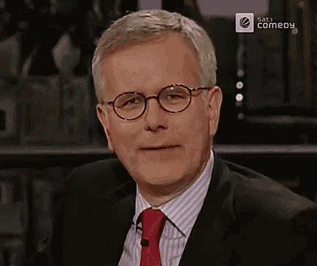 a man wearing glasses and a red tie is sitting in front of a screen that says saturn comedy