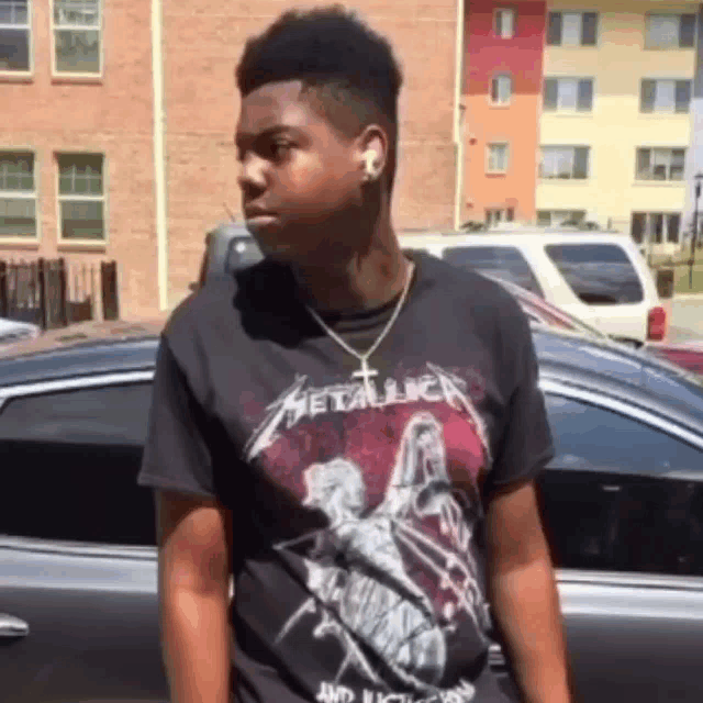 a young man wearing a black metallica t-shirt stands in front of a car .