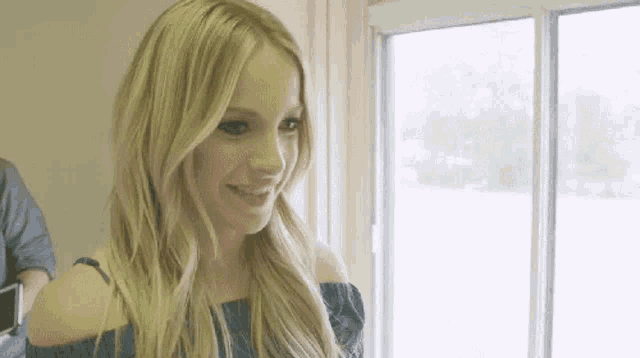 a woman in a blue off the shoulder top is smiling in front of a window .