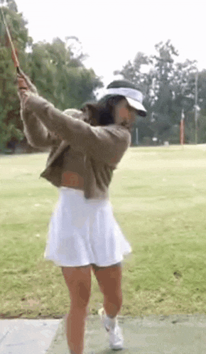 a woman in a white skirt and hat is swinging a golf club .