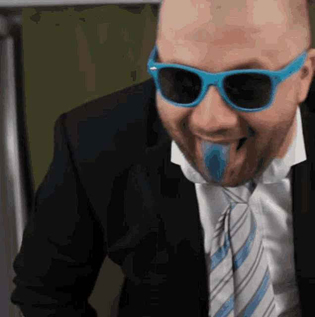 a man wearing sunglasses and a suit has a blue object in his mouth