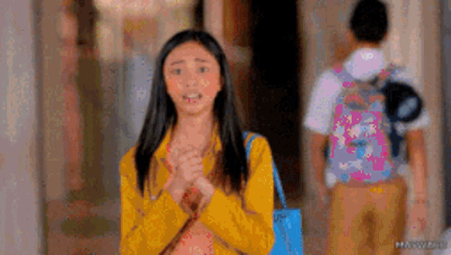 a girl in a yellow jacket is standing in a hallway with her hands folded in front of a man with a backpack .