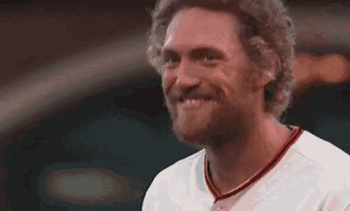 a man with a beard and curly hair is smiling and wearing a white baseball uniform .