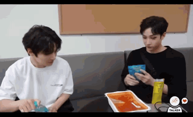 two young men are sitting on a couch looking at a container of food .